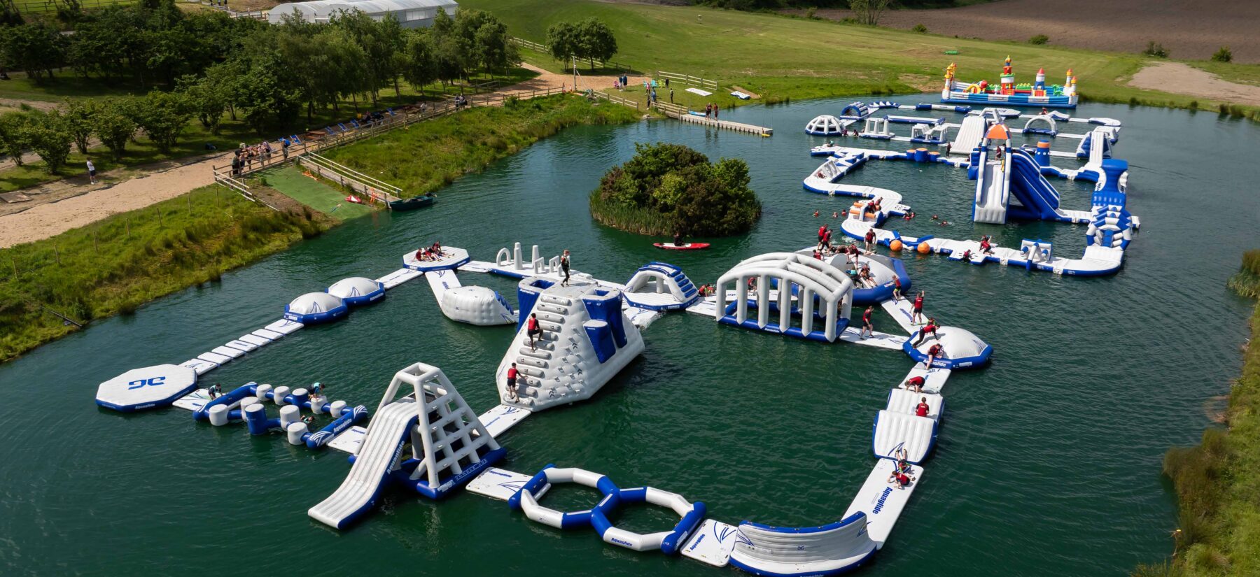 Aerial view of Flynn Park Adventure in Balbriggan, showing a large floating inflatable obstacle course on a lake with people climbing, sliding, and jumping between platforms.
