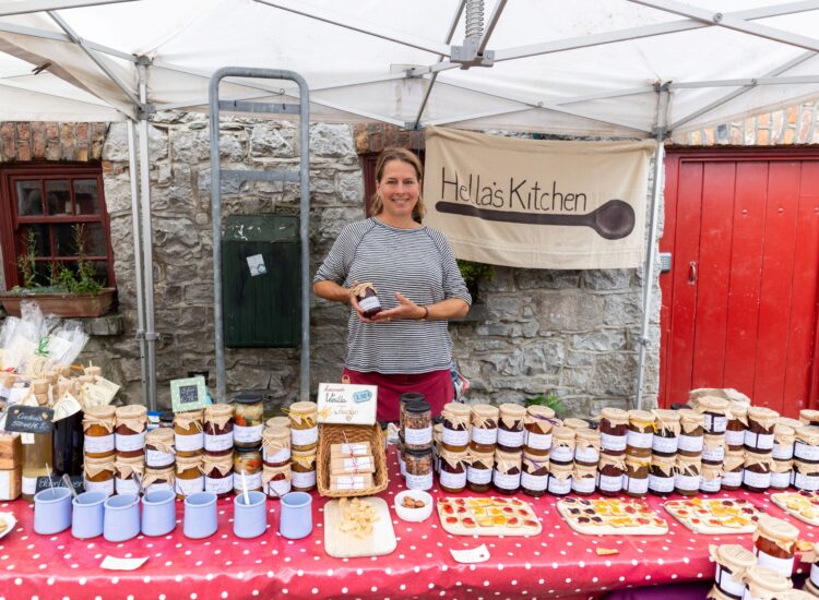 A market vendor standing behind a stall filled with jars of homemade jams, chutneys, and preserves under a canopy at Skerries Mills Farmers Market, with a sign reading “Hella’s Kitchen” hanging behind her.