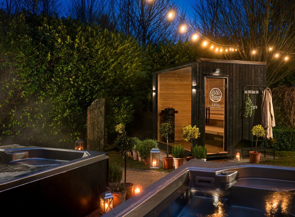 Two outdoor hot tubs steaming at night next to a wooden sauna building lit by warm string lights and lanterns.