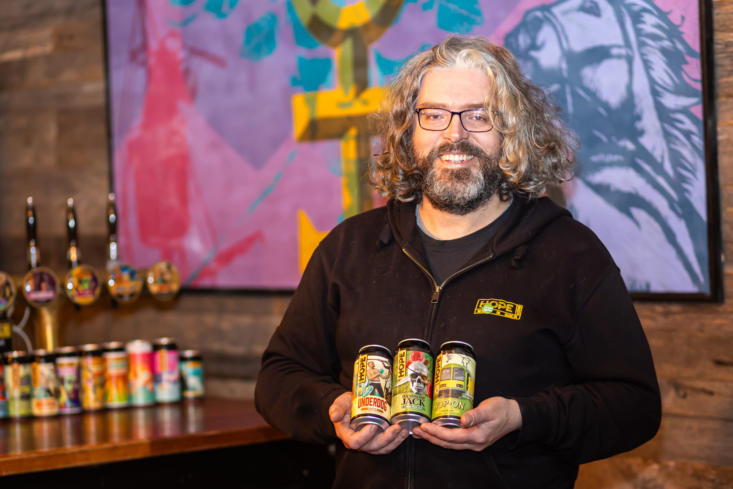 Richie Hamilton smiling while he holds three colourful Hope Beer cans inside a brewery.