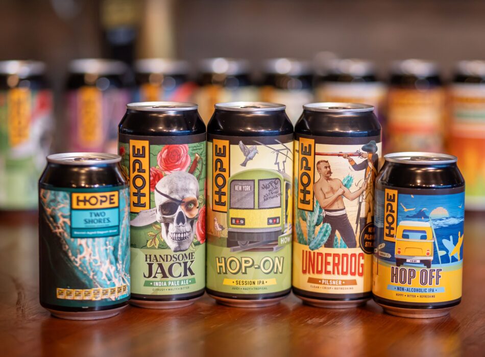 Five colurful artisanal-style Hope Beer cans lined up on a wooden counter.