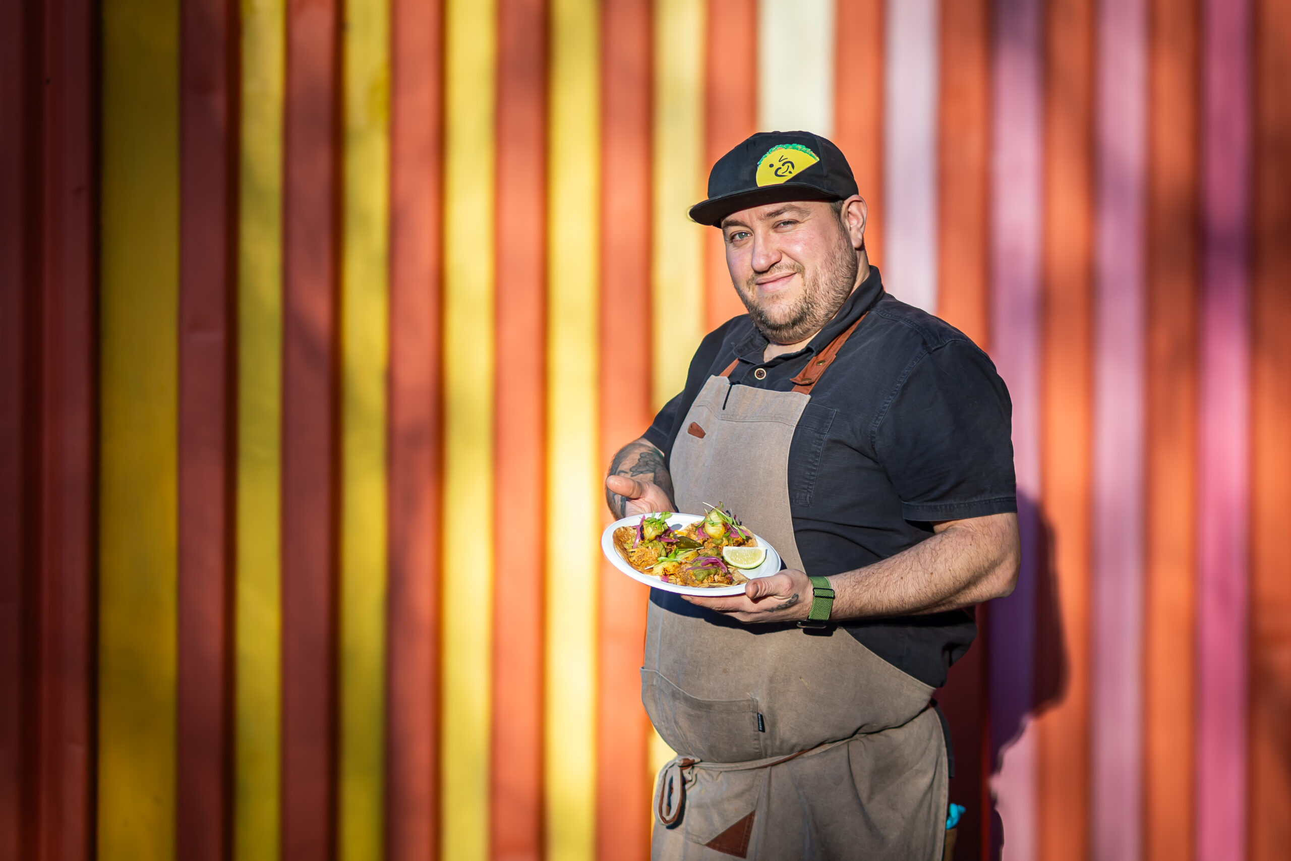 Jeremy Cuevas of La Cocina Cuevas posing with cusine infront of a colourful background.