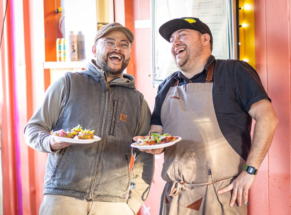 Two men standing side-by-side outdoors, laughing and holding small plates of tacos.