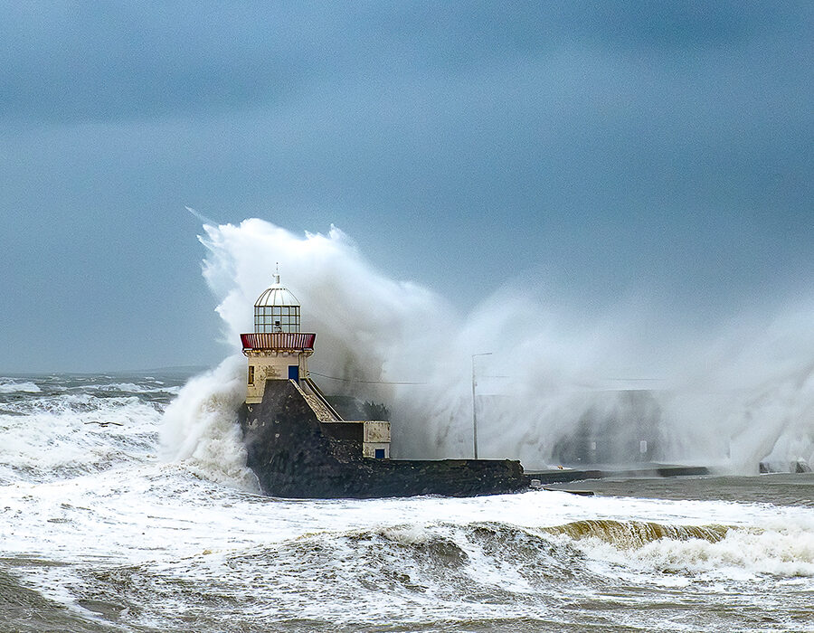 Large waves crash dramatically against a lighthouse and harbour wall during a storm, with dark skies and rough seas surrounding the scene.