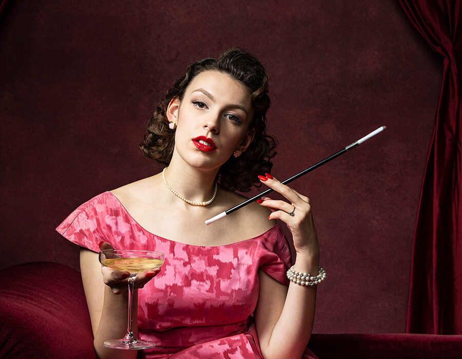 A woman in a vintage pink off the shoulder dress holding a coupe glass and a long cigarette holder.