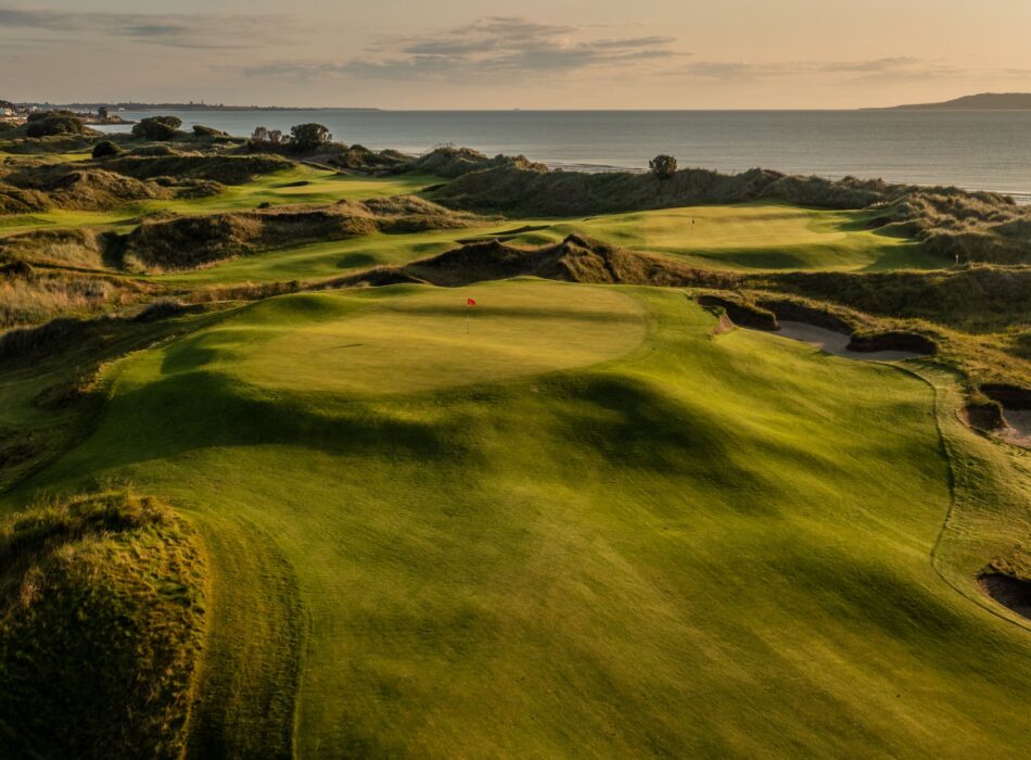 A fairway nestled in sandy dunes, looking back towards a large white resort building.