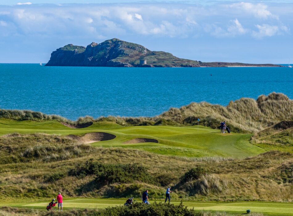 A coastal golf course green and tee box overlooking bright blue sea water, with a prominent rocky island featuring a historic Martello tower visable.