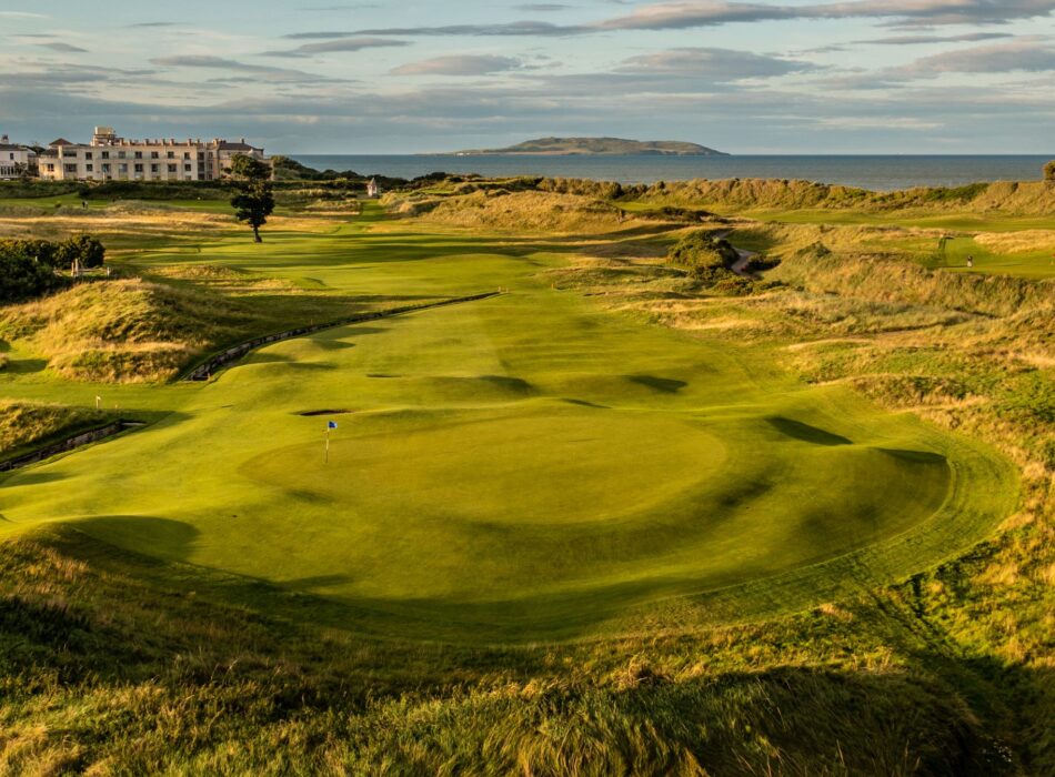 A fairway nestled in sandy dunes, looking back towards a large white resort building.