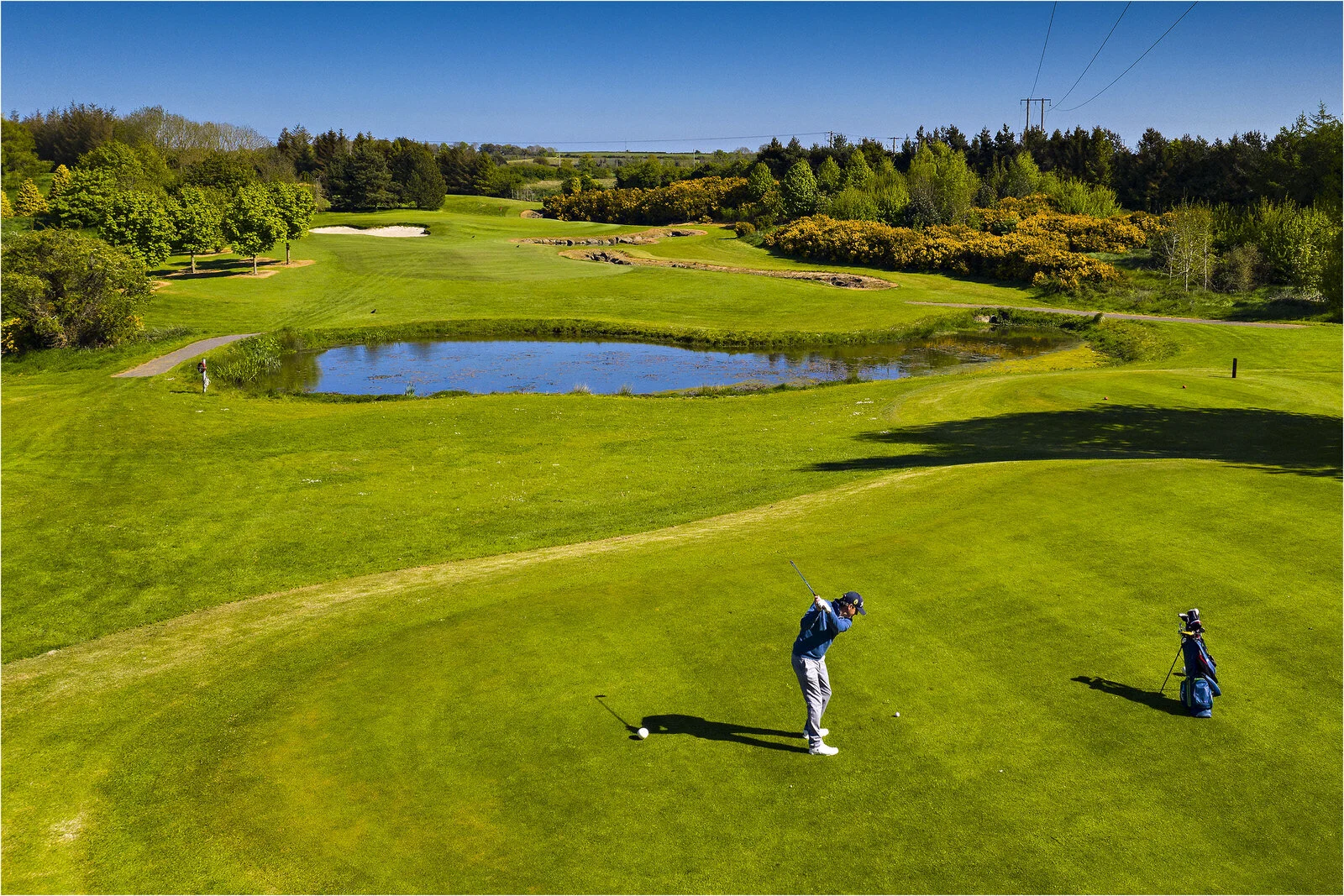 An aerial view of Balbriggan Golf Club on a bright summer day. A golfer prepares to tee off on a lush fairway overlooking a scenic course with rolling greens, sand bunkers, and a reflective lake. Mature trees and colourful gorse line the fairways, highlighting the attractive parkland setting.