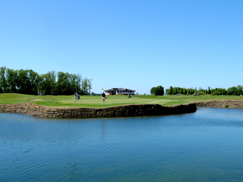 Golfers stand on a well-maintained green surrounded by water at Castleknock Golf Club on a bright summer day. The green sits on a stone-edged peninsula extending into a lake, creating a striking feature hole, while fairways, mature trees, and the clubhouse can be seen in the background beneath a clear blue sky.