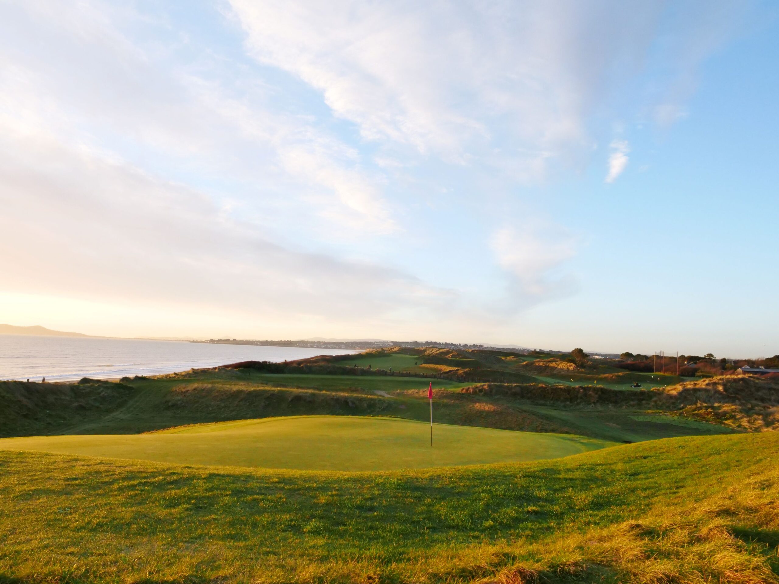 A coastal golf course at sunset, with a smooth putting green and red flag in the foreground surrounded by rolling links dunes. The sea stretches along the left-hand side, while warm golden light illuminates the fairways and dramatic sky, showcasing the natural beauty of Corballis Links Golf Course.