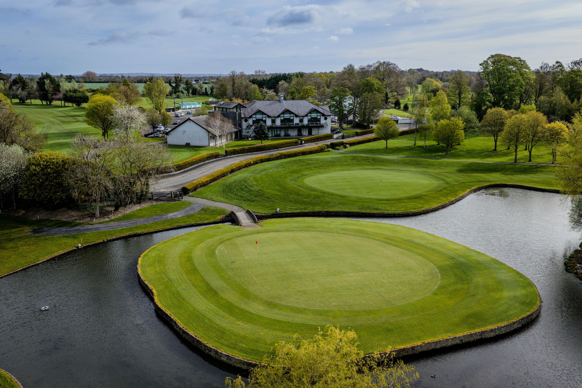 Corrstown golf course featuring a prominent, circular island green completely surrounded by calm water, with a small stone bridge connecting the island back to the main fairway.