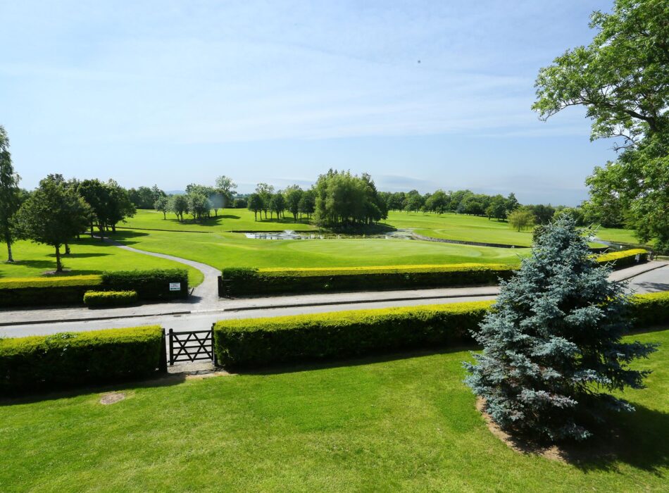 A panoramic view across Corrstown Golf Club, featuring immaculate fairways, a manicured green, mature trees, and a tranquil water feature under blue skies. The peaceful parkland setting showcases the beautifully maintained course, just a short distance from Dublin Airport.