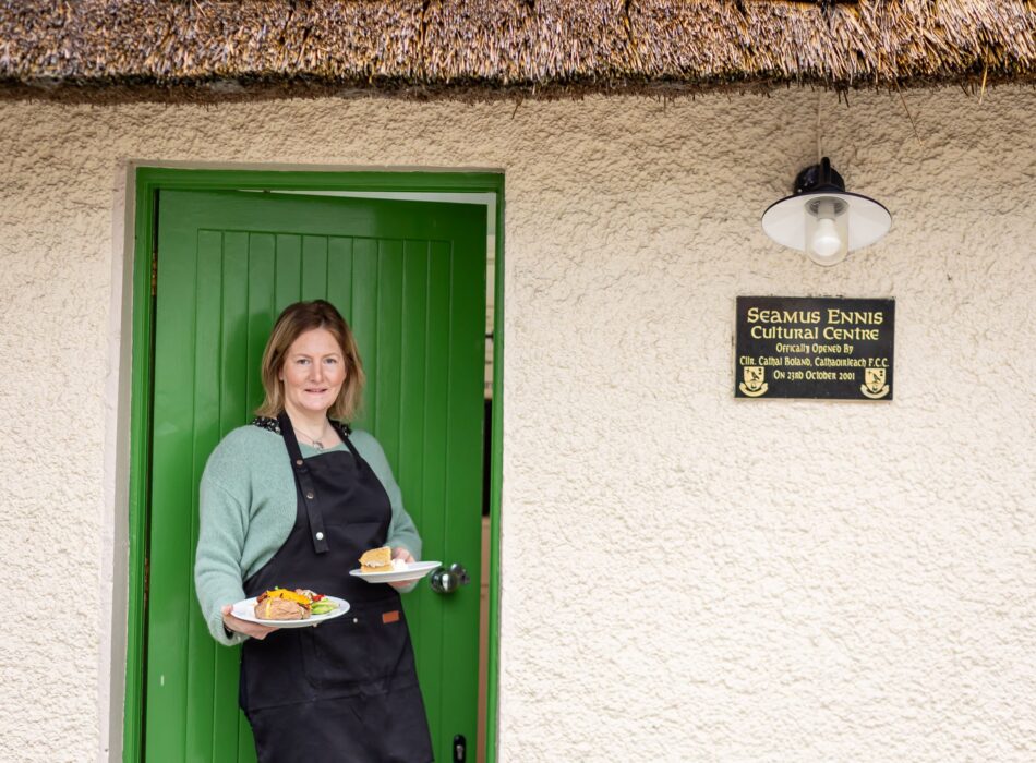 A woman in a black apron stands in the green doorway of a thatched-roof building, holding plates of food next to a commemorative plaque for the Seamus Ennis Cultural Centre.