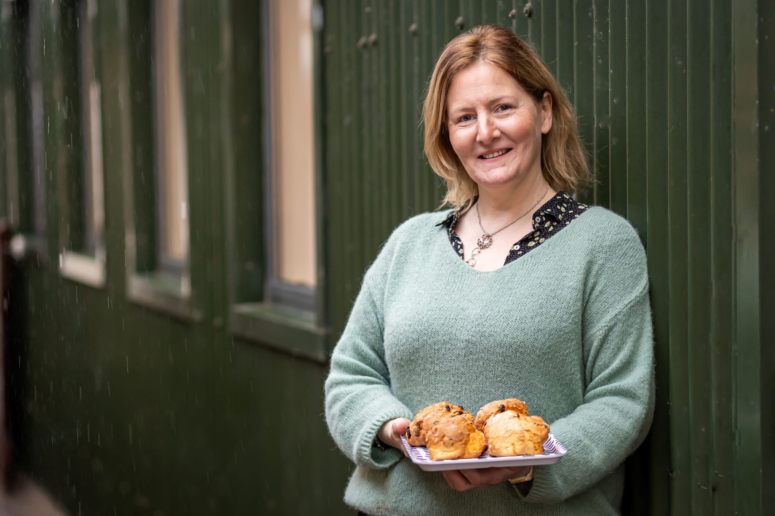 A smiling woman with blonde hair, wearing a light green knit sweater, leans against a green corrugated wall while holding a small tray of freshly baked scones.