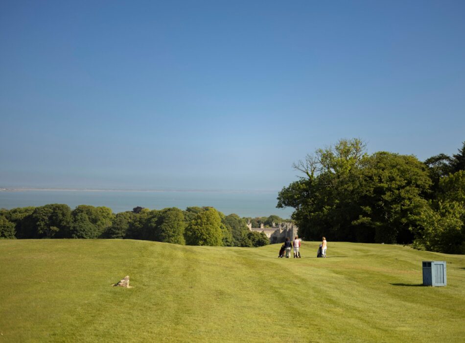 Golfers stand on the fairway of Deer Park Golf Course overlooking the grounds of Malahide Castle, with mature woodland, coastal views across Dublin Bay, and rolling green fairways stretching towards the sea.