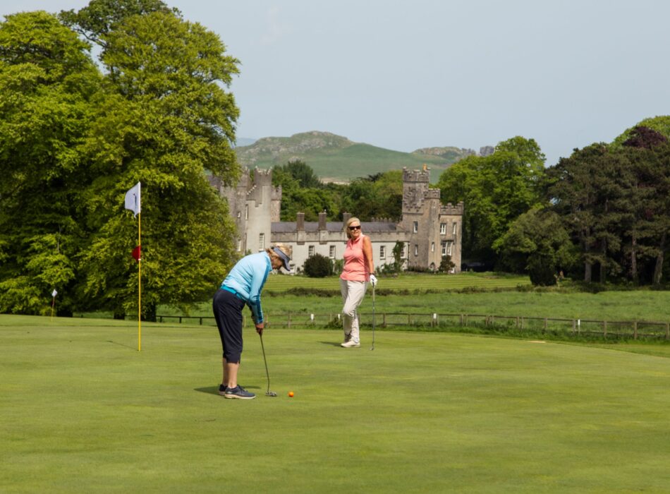 Two golfers on the fairway of Deer Park Golf Course overlooking the grounds of Malahide Castle, with mature woodland, coastal views across Dublin Bay, and rolling green fairways stretching towards the sea.