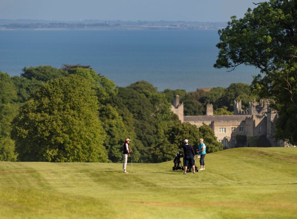 Golfers stand on the fairway of Deer Park Golf Course overlooking the grounds of Malahide Castle, with mature woodland, coastal views across Dublin Bay, and rolling green fairways stretching towards the sea.