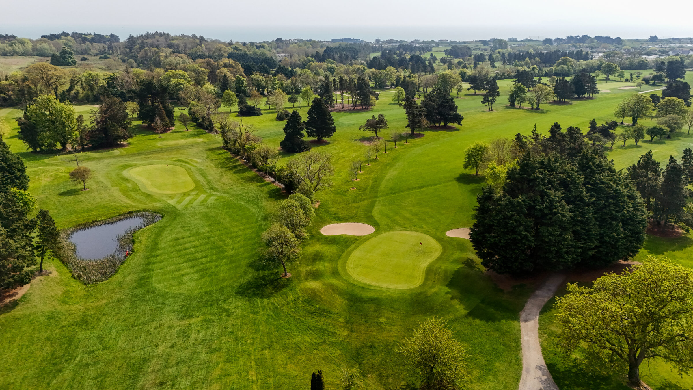 An aerial view of Donabate Golf Club showcasing lush fairways, manicured greens, mature woodland, and a tranquil water feature. The expansive parkland course offers a scenic and enjoyable golfing experience in the heart of Fingal, just minutes from Dublin Airport and the coast.