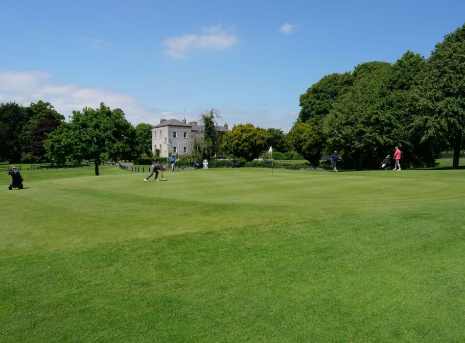 A wide landscape view of Elmgreen Golf Club under a clear blue sky with light clouds. In the background, several people are playing golf near a flag stick and a historic stone building stands next to a line of mature green trees.