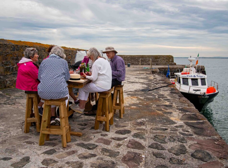A group of adults sitting at a wooden picnic table with barstools on a stone pier next to the ocean, with a boat in the background.