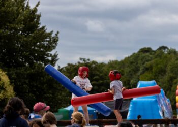Two children wearing protective headgear playfully jousting with large inflatable batons, one blue and one red.