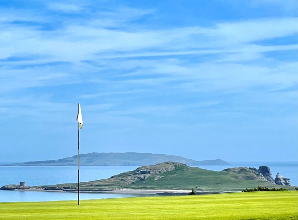 A flag stick stands in the against calm blue waters. In the distance, the coastline and the Wicklow Mountains create a dramatic backdrop, highlighting the unique coastal setting of the course.