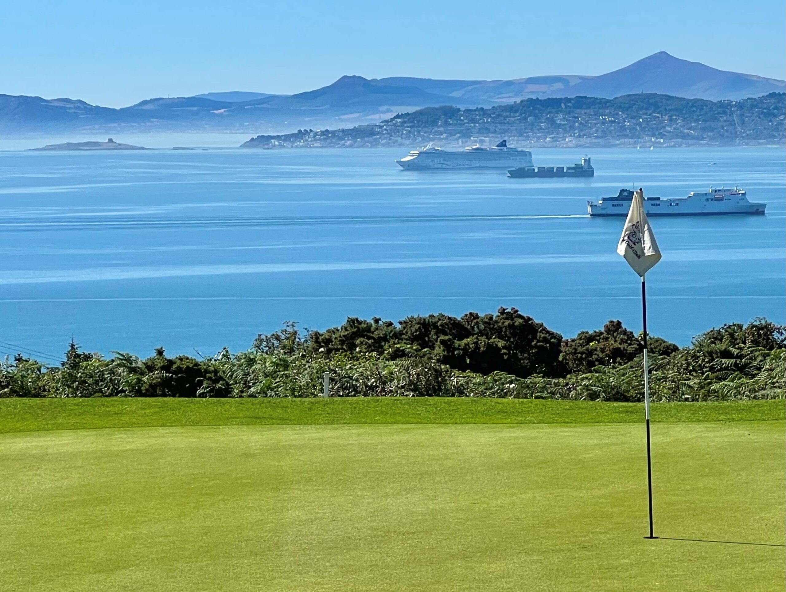 A pristine golf green at Howth Golf Club overlooks Dublin Bay on a clear day. A flagstick stands in the foreground, while ferries, a cruise ship, and cargo vessels cross the calm blue waters. In the distance, the coastline and the Wicklow Mountains create a dramatic backdrop, highlighting the unique coastal setting of the course.