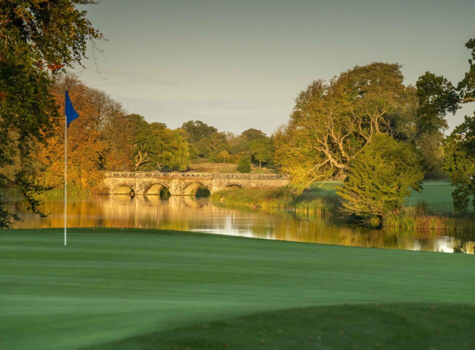 A tranquil golf green at Luttrellstown Castle Golf Club beside a reflective lake, with a blue flag marking the hole in the foreground. A historic stone bridge arches across the water in the background, framed by mature trees in warm autumn colours. The calm water and golden evening light create an elegant and picturesque parkland setting.