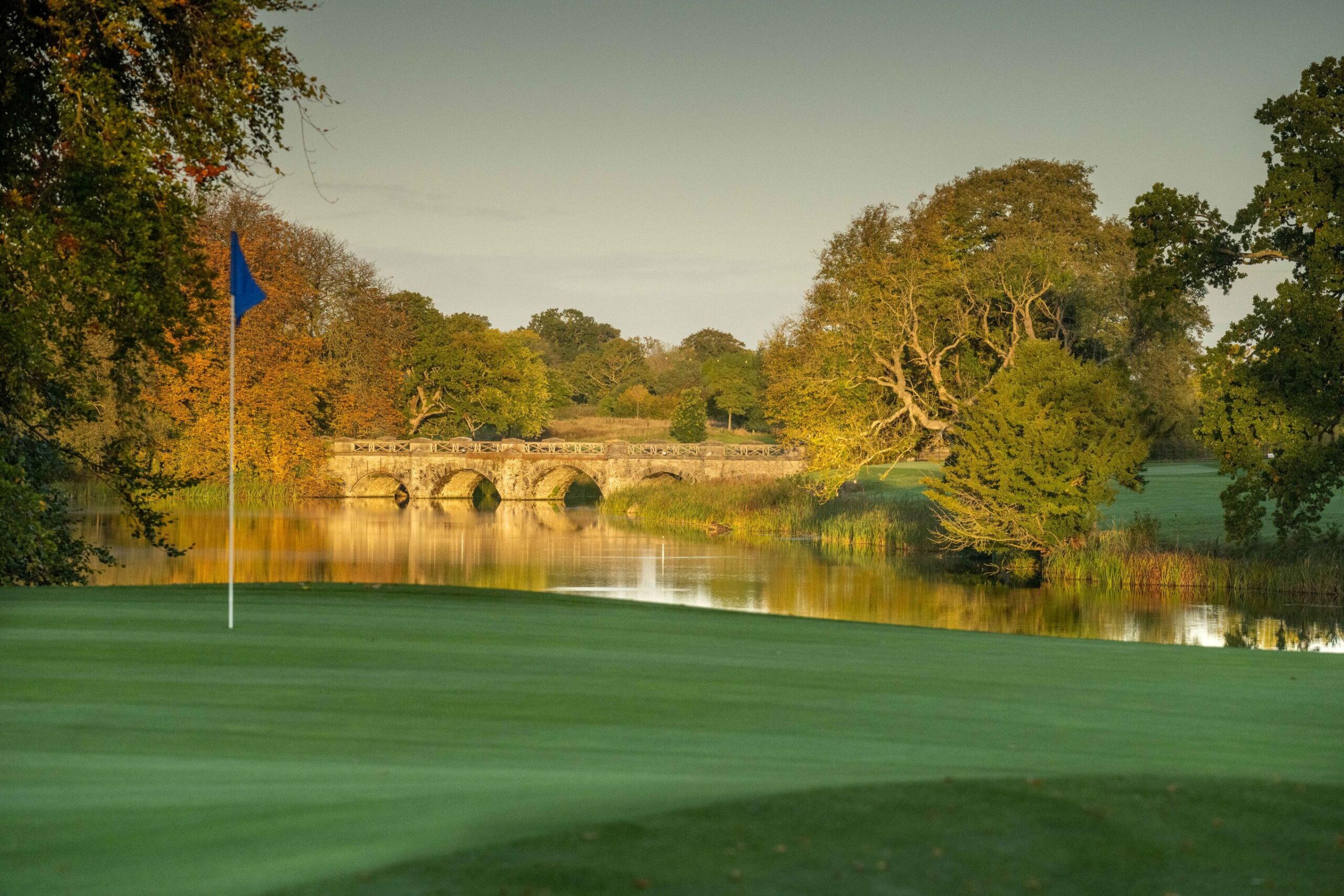 A tranquil golf green at Luttrellstown Castle Golf Club beside a reflective lake, with a blue flag marking the hole in the foreground. A historic stone bridge arches across the water in the background, framed by mature trees in warm autumn colours. The calm water and golden evening light create an elegant and picturesque parkland setting.
