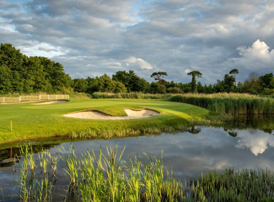 A manicured golf green with deep sand bunker sitting directly behind a calm water hazard, with reeds and white clouds reflecting the water's surface.