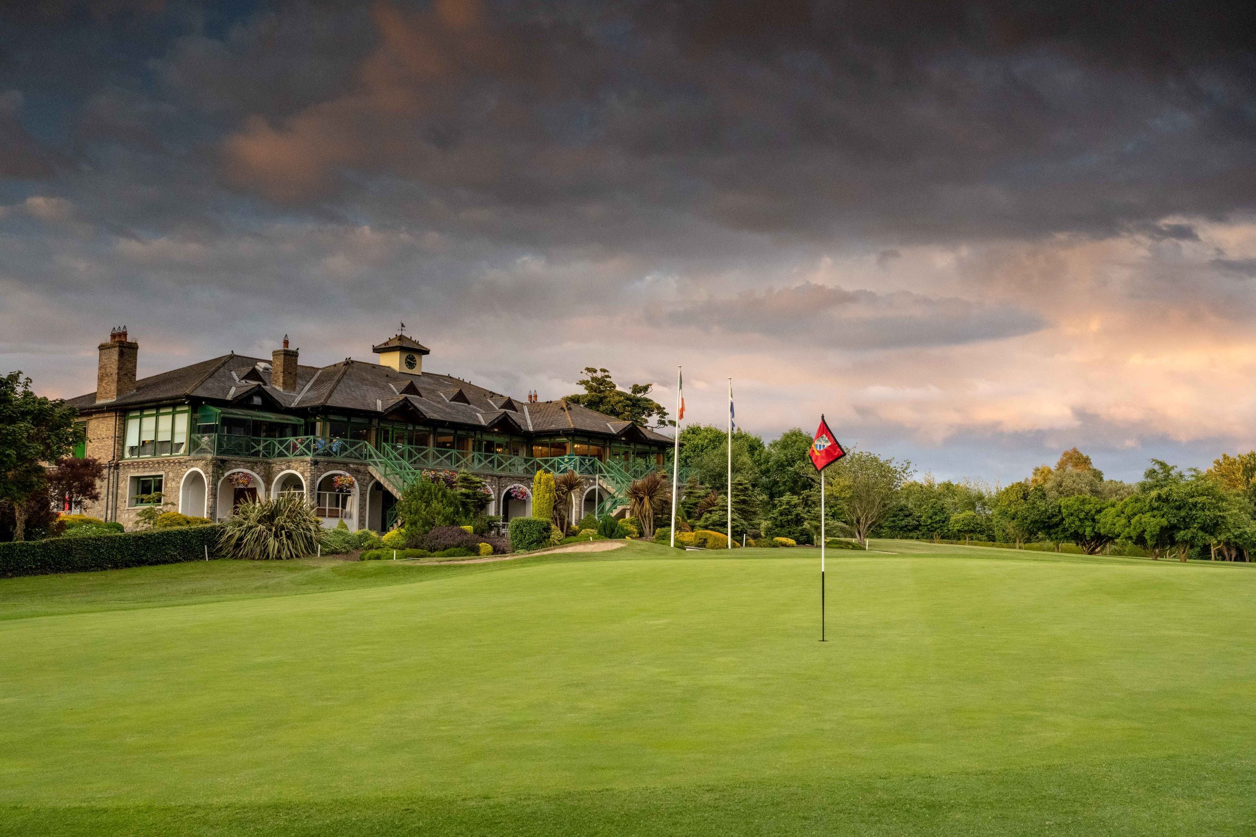 Landscape shot of Malahide Golf Club during a vibrant sunset. A large and historic looking stone clubhouse overlooks a lush, manicured green golf course under a sunset of grey and orange clouds.