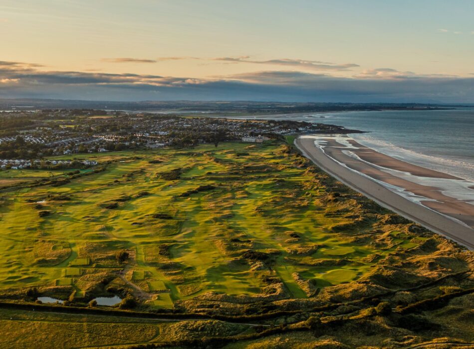 A high-altitude aerial view of an expansive green links golf course stretching along a wide sandy coastline and the sea under a warm sunny sky.