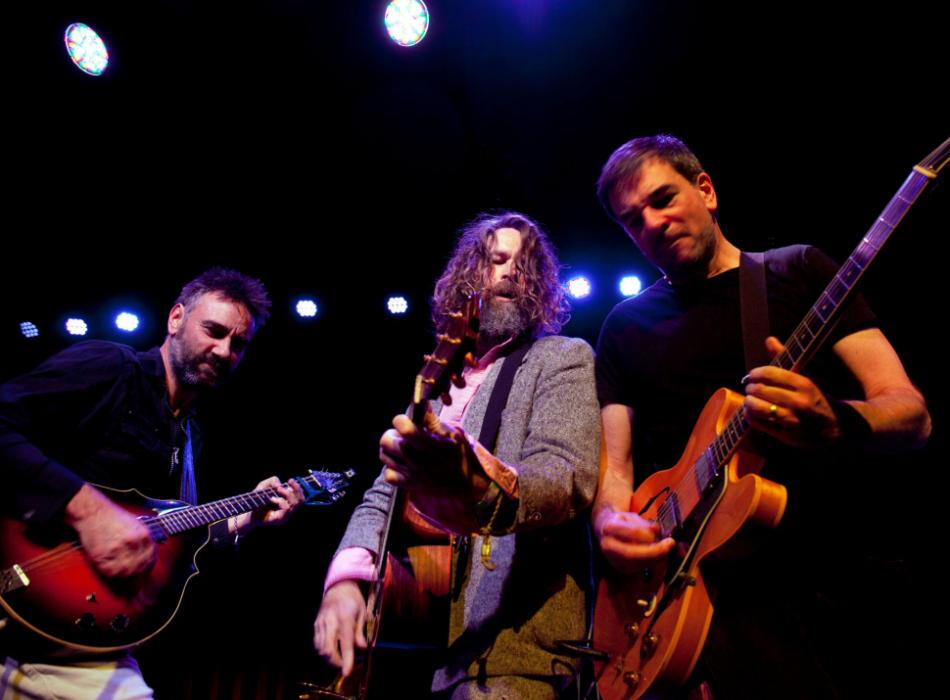 Three male musicians performing live on stage under stage lighting. The musician on the left plays a mandolin, the centre musician plays an acoustic guitar and the last musician on the right plays an orange electric guitar.