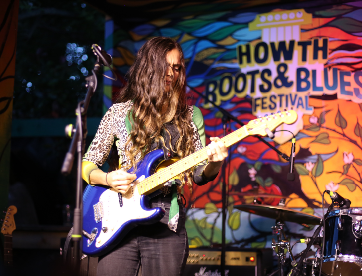 A female musician with long, wavy brown hair performs live on stage, playing a vibrant blue electric guitar. She wears a leopard-print long-sleeve top under a green vest and dark plaid trousers.