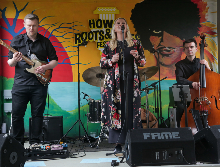 A musical band performs live on stage in front of a colourful mural backdrop featuring the “Howth Roots & Blues Festival” logo. A female vocalist in a long black dress with a red and white floral pattern sings into a microphone. Behind her, a man plays an electric guitar, while another musician plays a double bass in the foreground. Stage monitor are positions on the floor.
