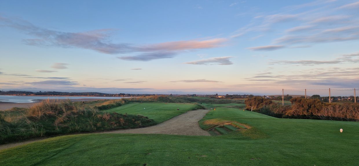 A landscape view of a links golf course at sunset. A dirt pathway cuts through the green lawns, leading toward a low dunes and coastal grass on the left. In the background a body of water sits beneath a large sky filled with soft blue and pink clouds.