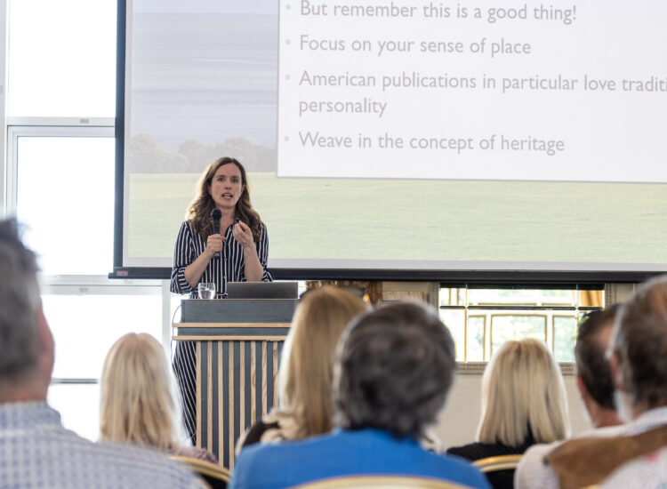Speaker presenting at a tourism conference, standing at a podium with a microphone in front of a projected presentation screen while an audience listens attentively.