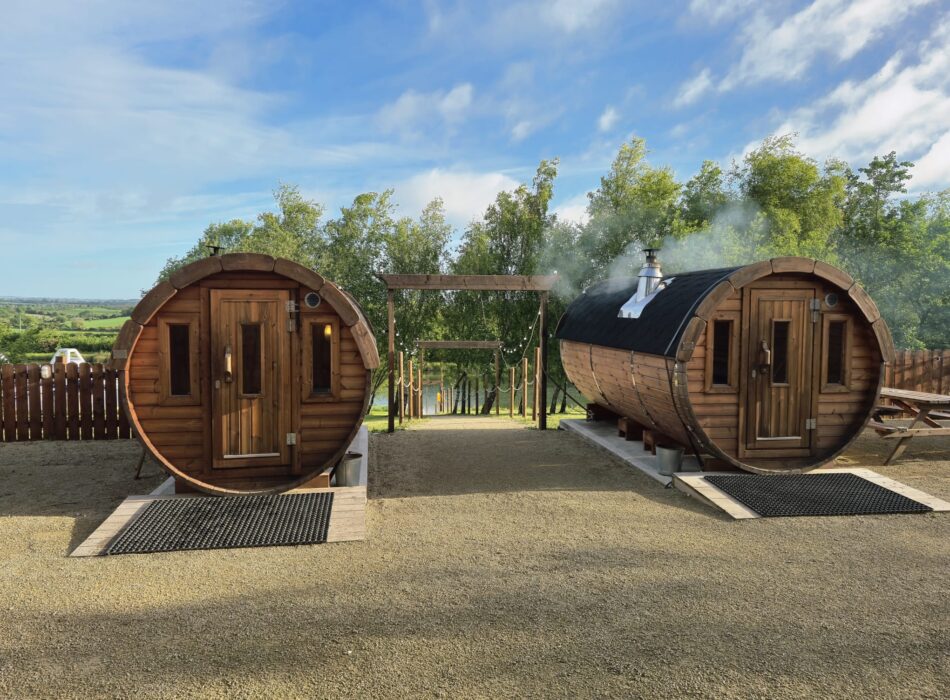 Two outdoor barrel saunas side-by-side with a wooden archway leading to trees and water under a blue sky.