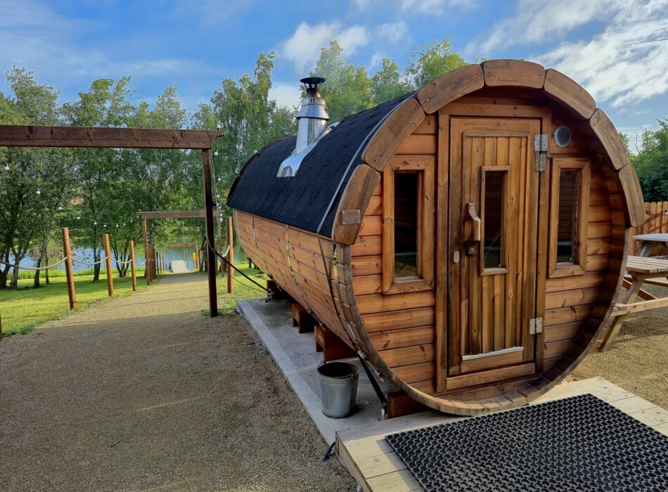 Exterior side view of a wooden barrel sauna with a black rood and silver chimney outdoors.