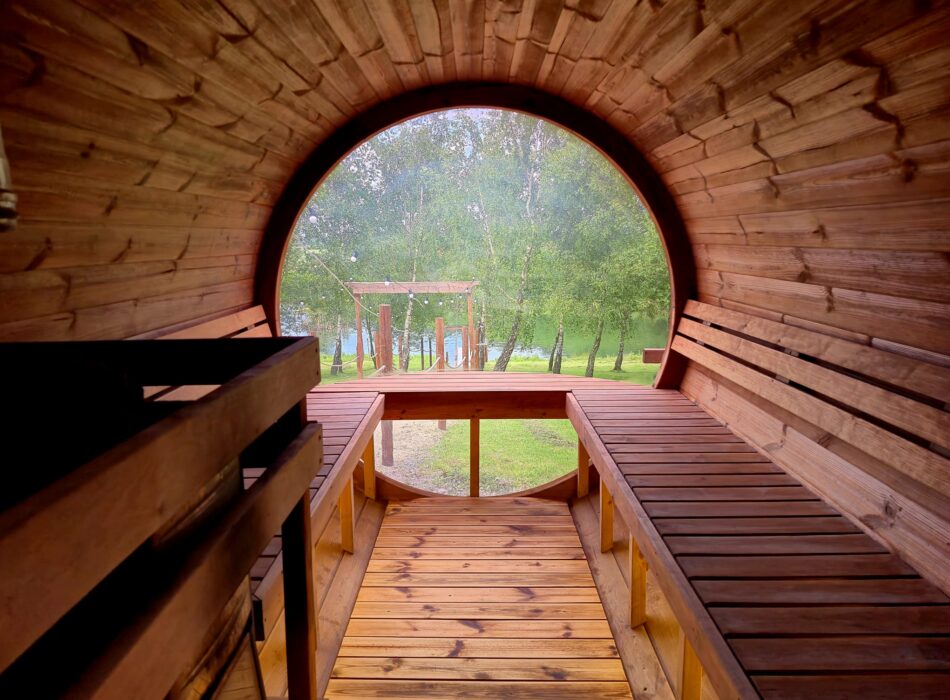 Interior view a wooden barrel sauna with slatted benches looking out a large circular window at a green landscape.