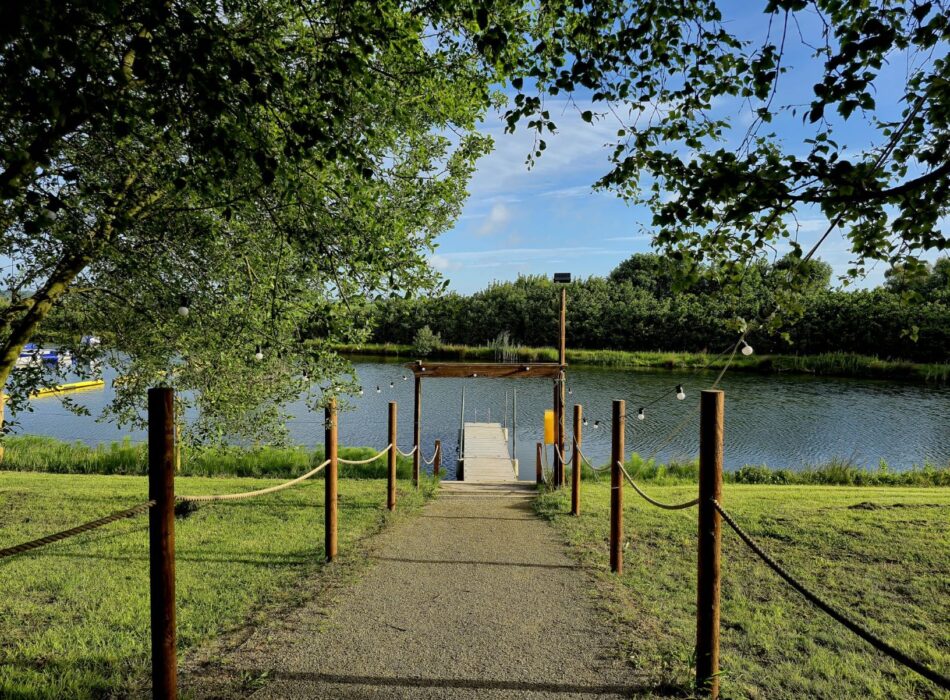 A rope-lined gravel path leading down to a wooden archway and dock on a calm lake.