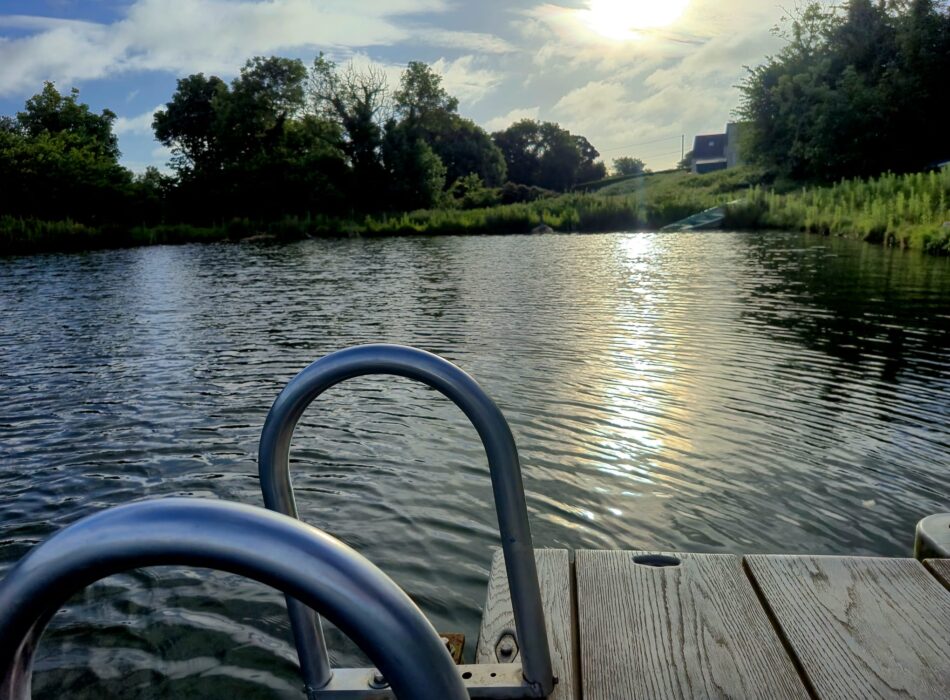 A calm lake pictured with a wooden deck and a metal ladder off the deck.