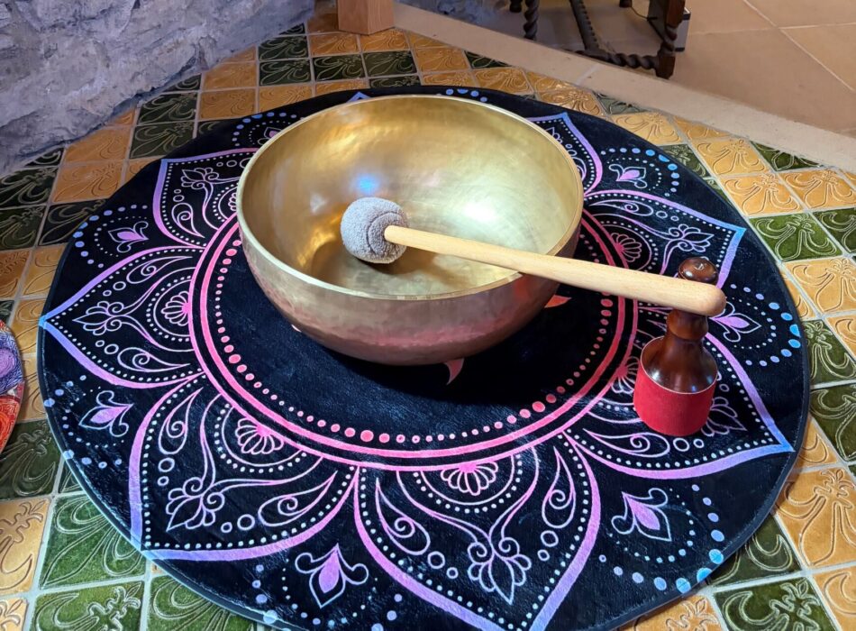 A brass singing bowl with a wooden mallet resting inside it, placed on a circular mandala mat on a tiled floor.