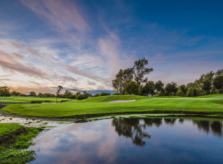 A green golf fairway and bunker reflecting perfectly into a calm body of water in the foreground, set against trees and a vibrant pink and blue twilight.