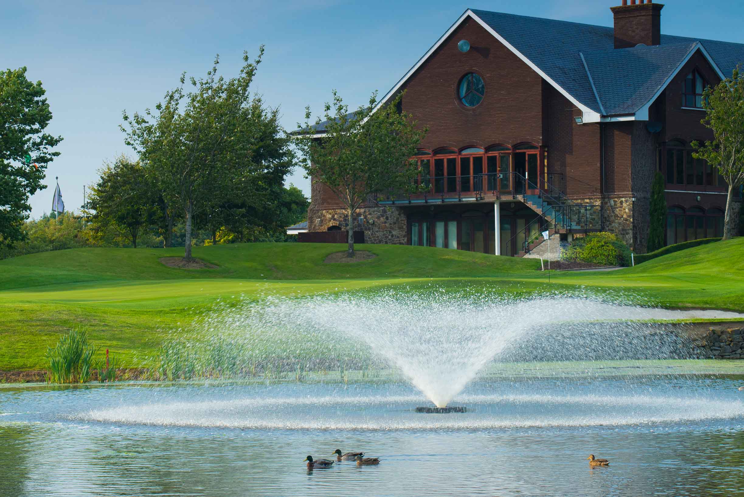 A large red brick and stone golf clubhouse building overlooking a water hazard feature with a spraying fountain and several ducks swimming on the surface.
