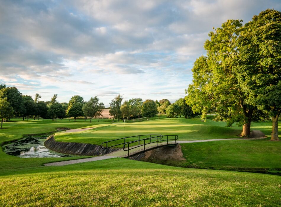 A sunlit parkland golf course with a black metal bridge crossing a winding stream separating lush green fairways.