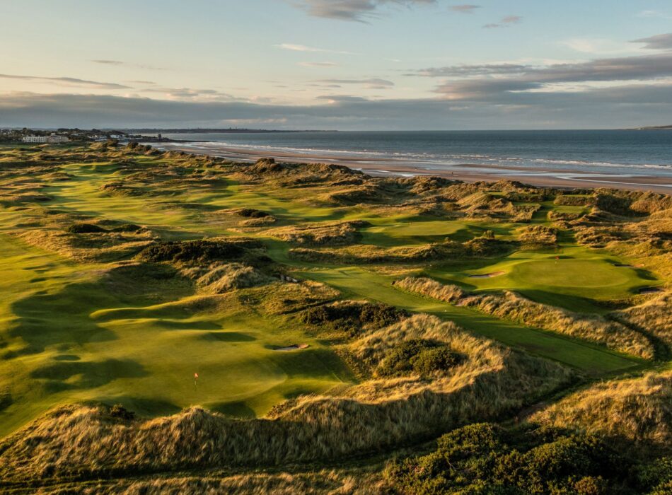 An aerial shot of green links golf course winding through rolling sand dunes alongside a wide sandy beach.