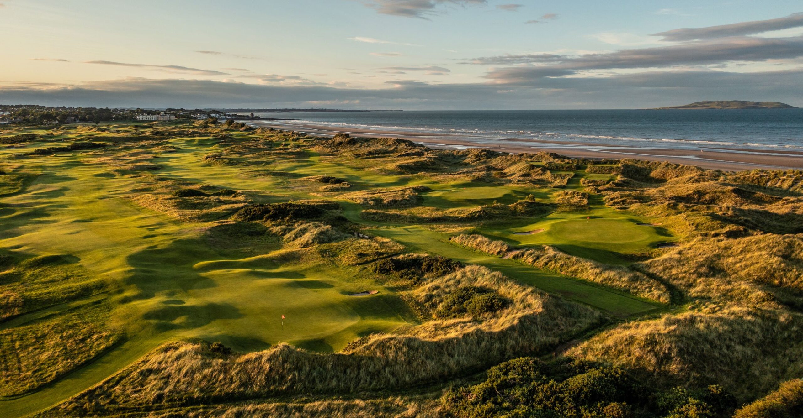 An aerial shot of green links golf course winding through rolling sand dunes alongside a wide sandy beach.