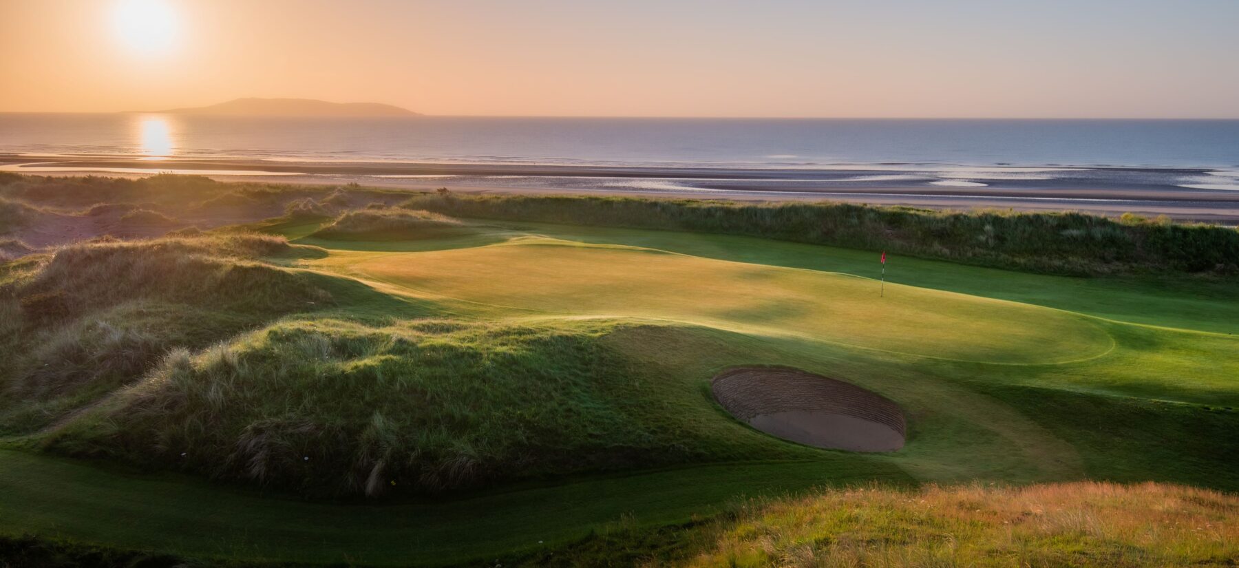 A stunning links golf course at sunrise, with a perfectly maintained green and bunker nestled among rolling sand dunes. The early morning sun reflects across the calm sea beyond the course, while golden light highlights the contours of the landscape, showcasing the dramatic coastal setting of The Island Golf Club.